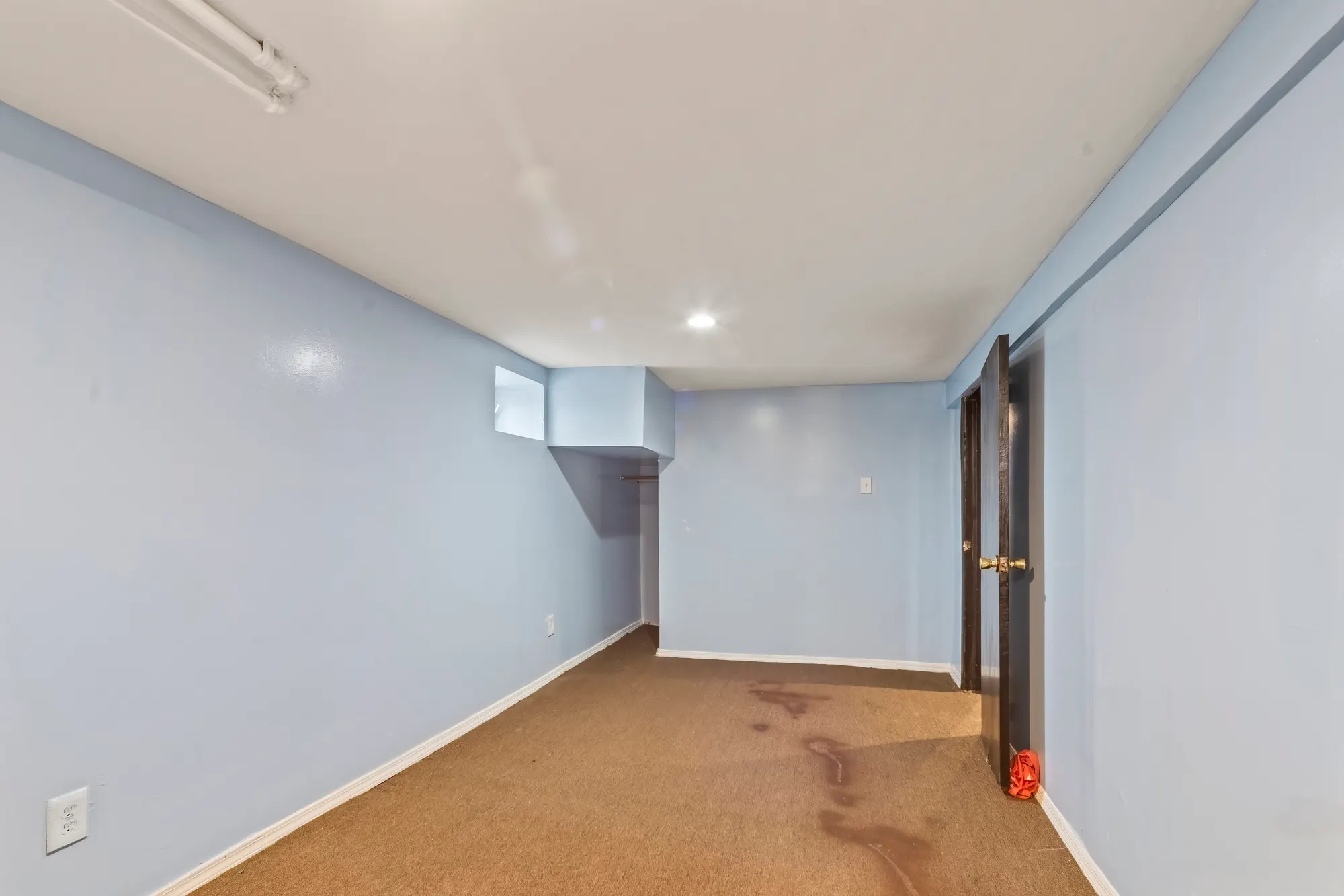Finished basement room in a Queens Village house with light blue walls, beige carpet, a fluorescent ceiling fixture and a dark door standing open