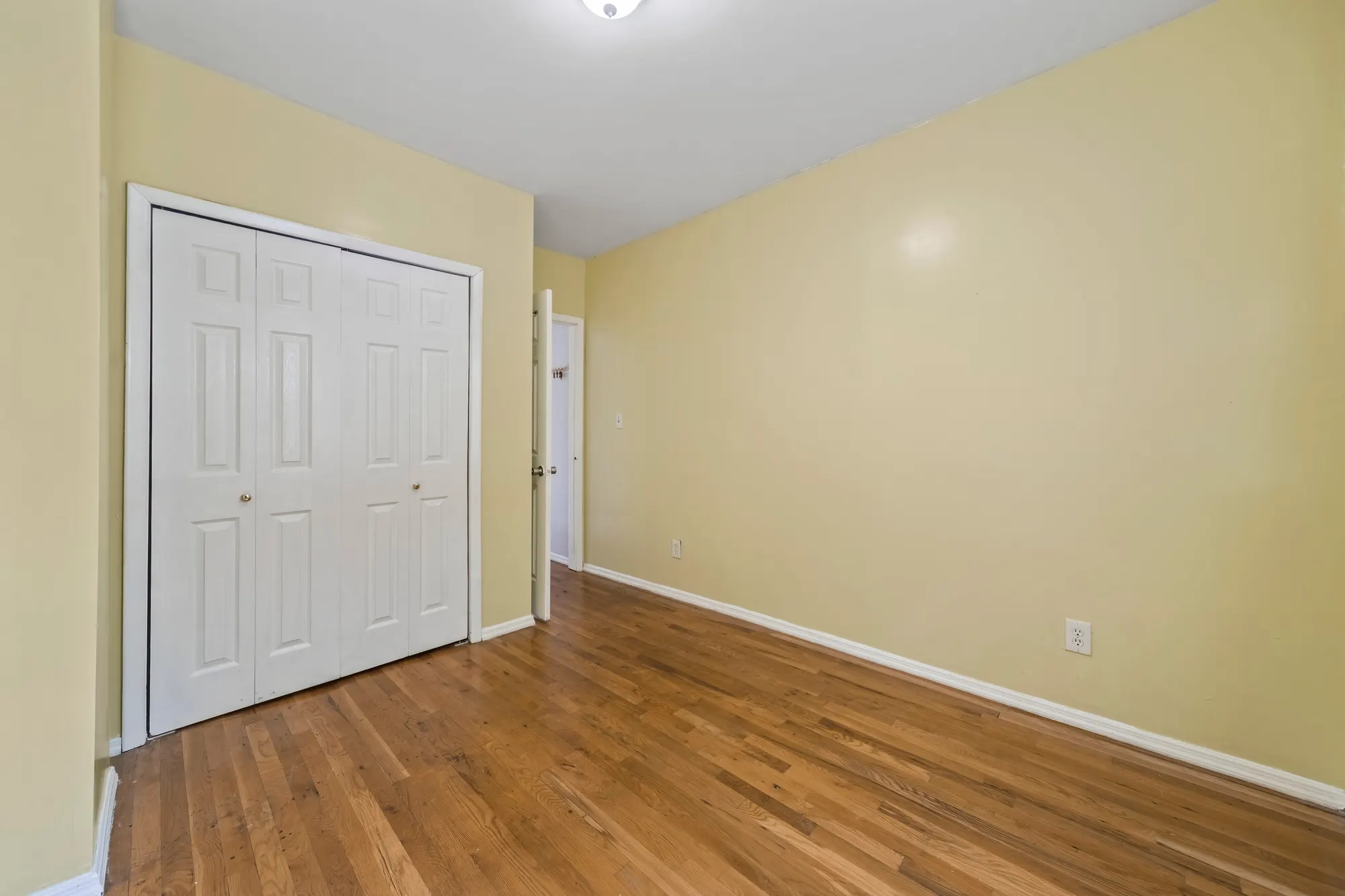 Empty bedroom in a Queens Village house with pale yellow walls, white louvered closet doors and refinished oak floors running to an open doorway