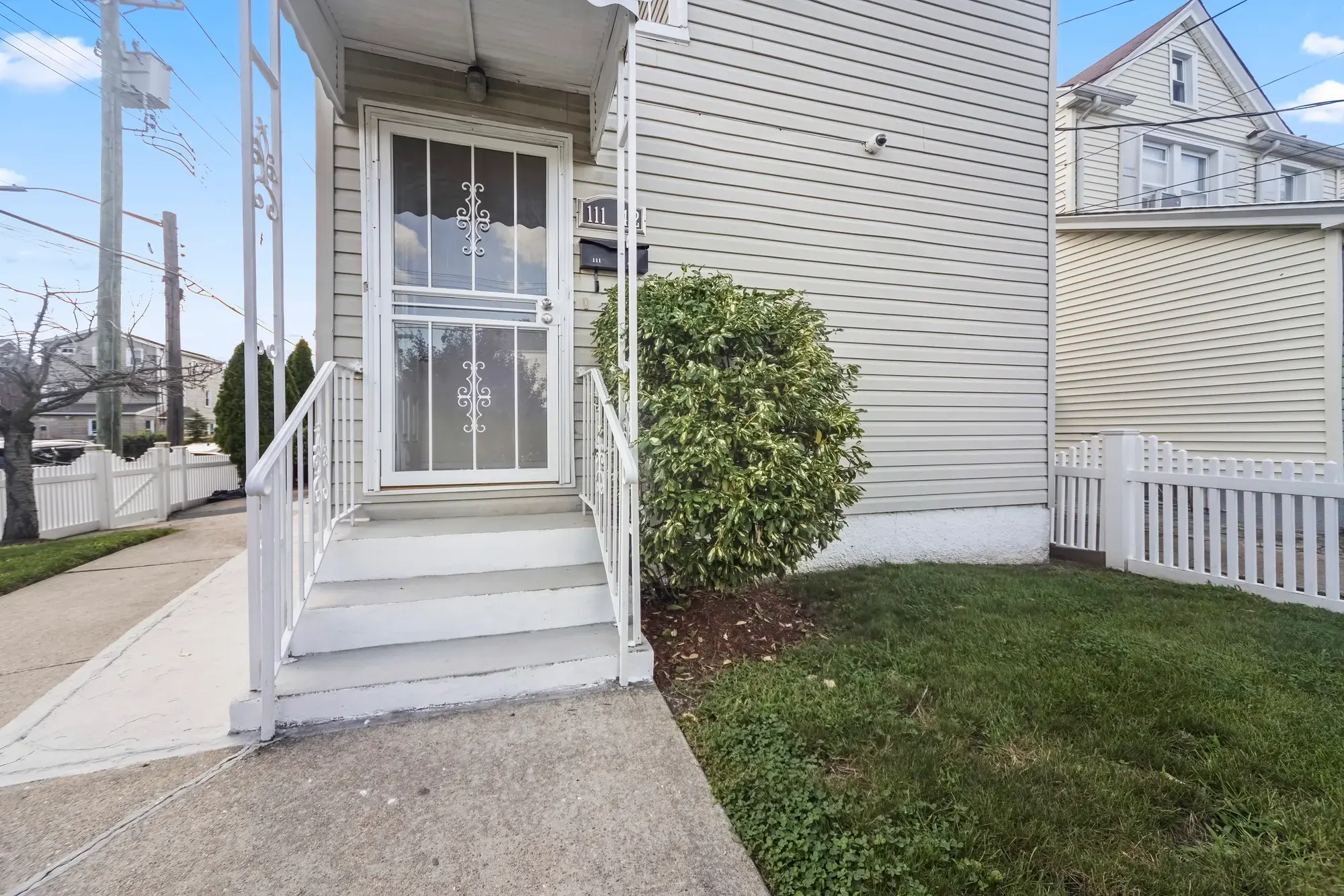 Side entrance of a Queens Village house, a storm door with a decorative screen at the top of four concrete steps with a white railing, a trimmed shrub beside the path and a white picket fence on the lawn