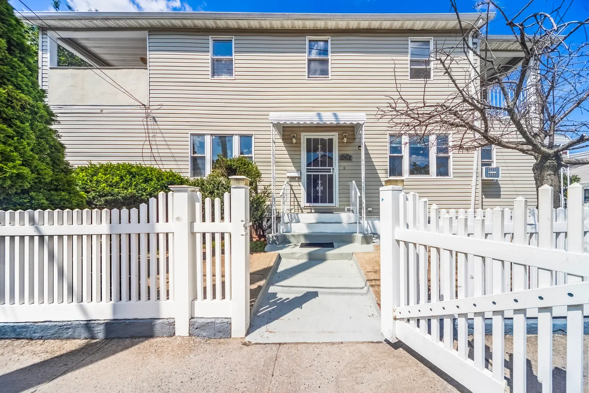 Front elevation of a two-story vinyl-sided house in Queens Village with a white picket fence, a concrete walk to the front steps, clipped hedges and a bare tree against a blue sky, shot for a listing by OpenDoors360
