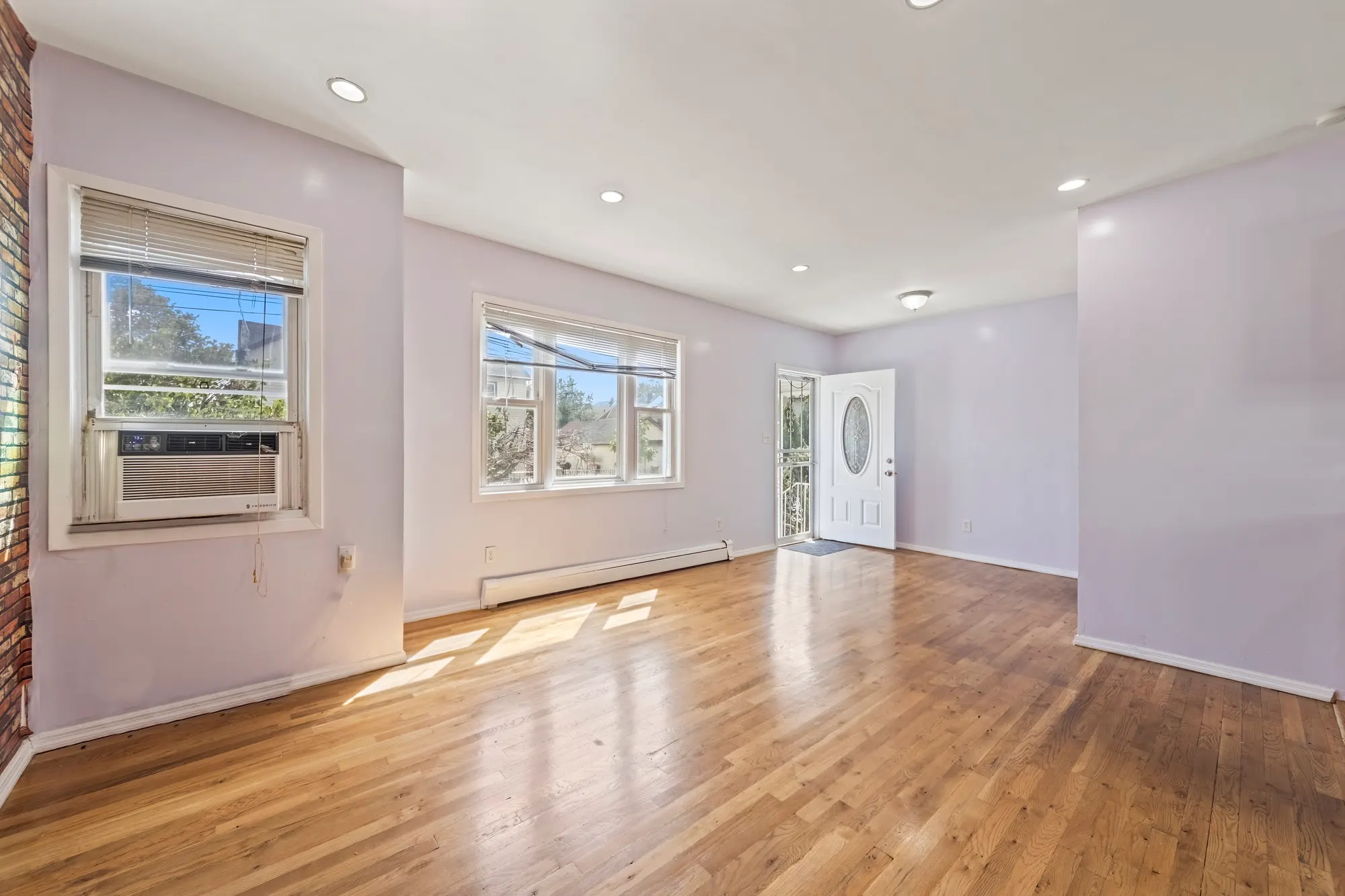 Empty living room in a Queens Village house with pale lilac walls, refinished oak floors, two windows with a window air conditioner, a baseboard heater and a white front door with an oval glass panel