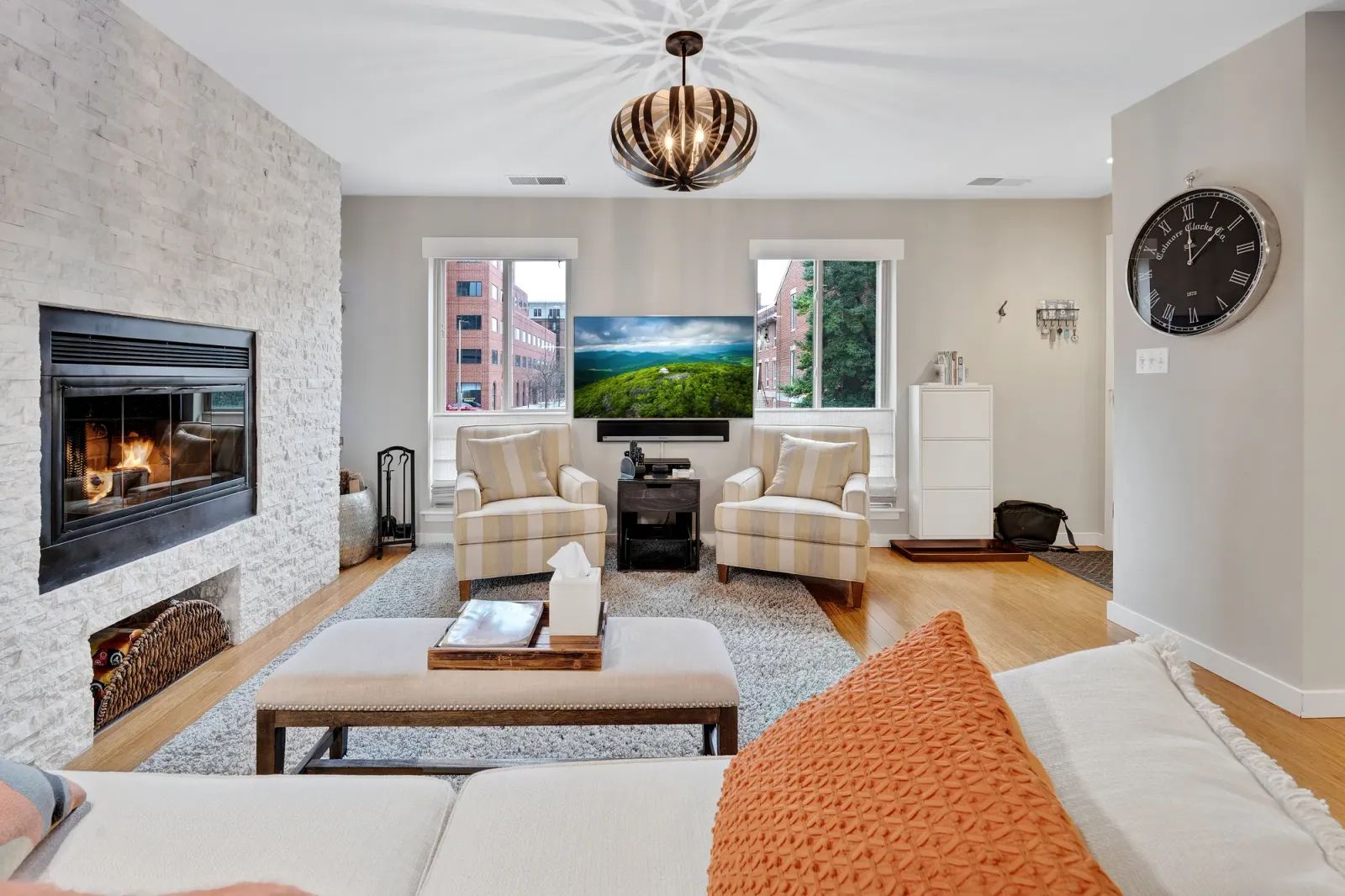 OpenDoors360 real estate photo of a living room with a white stacked-stone fireplace wall and the fire lit, two cream and tan striped armchairs flanking a wall-mounted TV showing a still green mountain image between two windows, a white tissue box and a wood tray on an upholstered ottoman, a white three-drawer file cabinet, a black bag on the floor by the doorway, keys hanging on a wall hook, a large round wall clock and a wire-globe pendant light