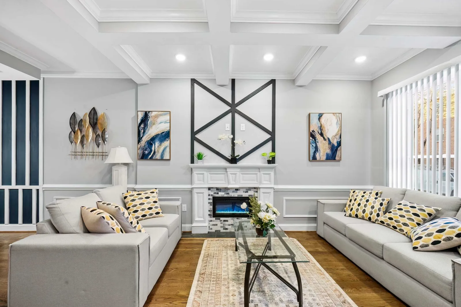 OpenDoors360 HDR real estate photography of a Baltimore, Maryland rowhouse living room shot straight on: two light gray sofas with yellow-and-black leaf pillows, a white coffered ceiling, a black geometric trim panel over a white fireplace mantel with mosaic tile, two blue-and-gold abstract paintings, a navy vertical-stripe accent wall at left, vertical blinds, a glass coffee table on a patterned rug and oak floors.