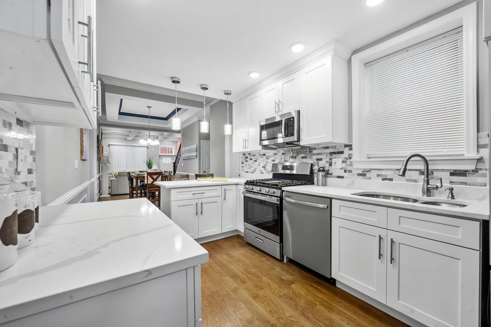 OpenDoors360 HDR real estate photography of a Baltimore, Maryland rowhouse kitchen: white shaker cabinets, a gray marble-pattern brick backsplash, white quartz counters, a stainless gas range, dishwasher and over-range microwave, three glass pendant lights, a window with white blinds over the sink and a sightline through to the dining table and coffered living room.