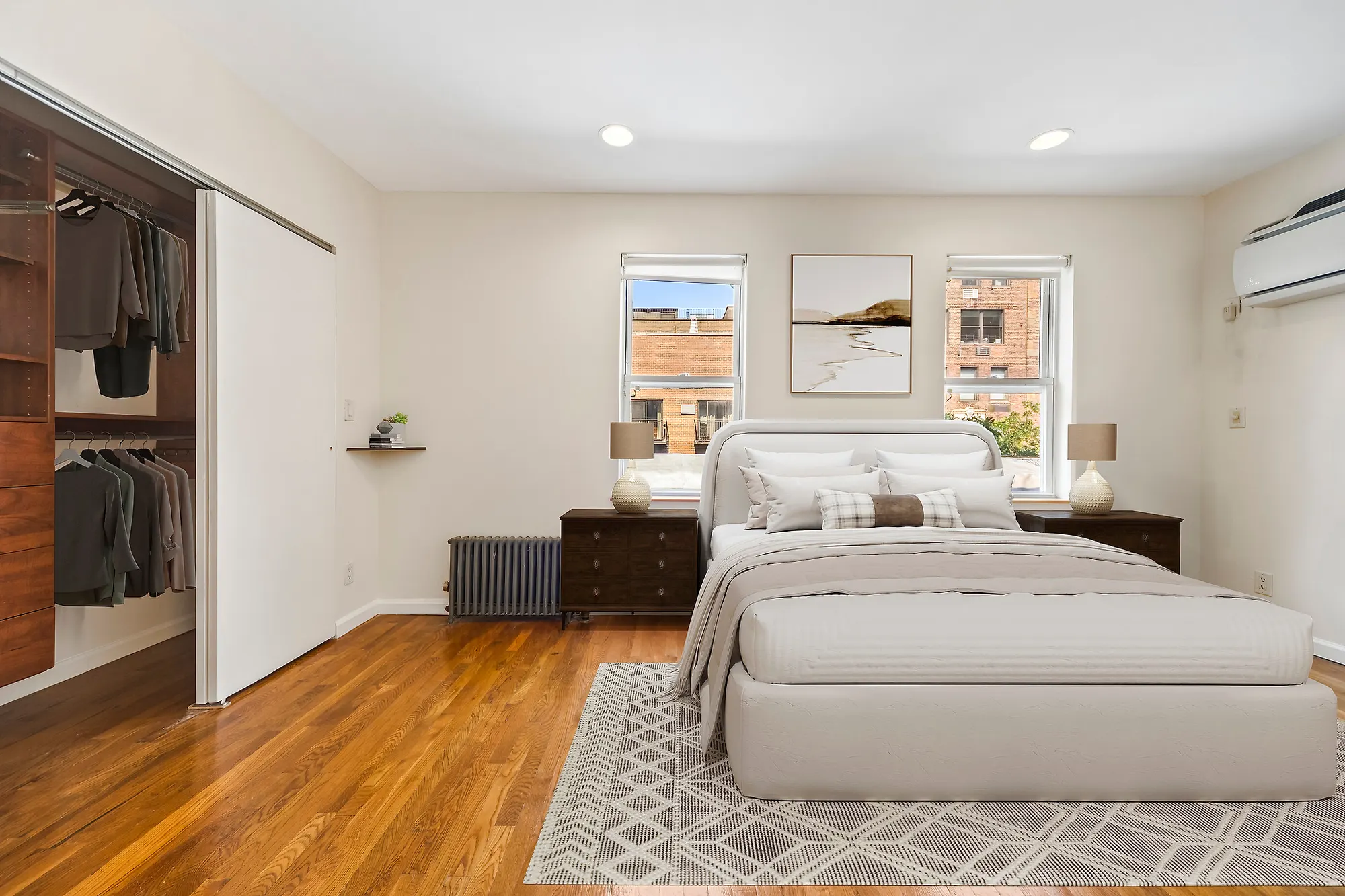 Virtually staged bedroom in an Upper East Side apartment, a bed with a cream upholstered headboard between two dark wood nightstands with lamps, a framed landscape above it, a patterned rug and an open closet fitted with hanging clothes