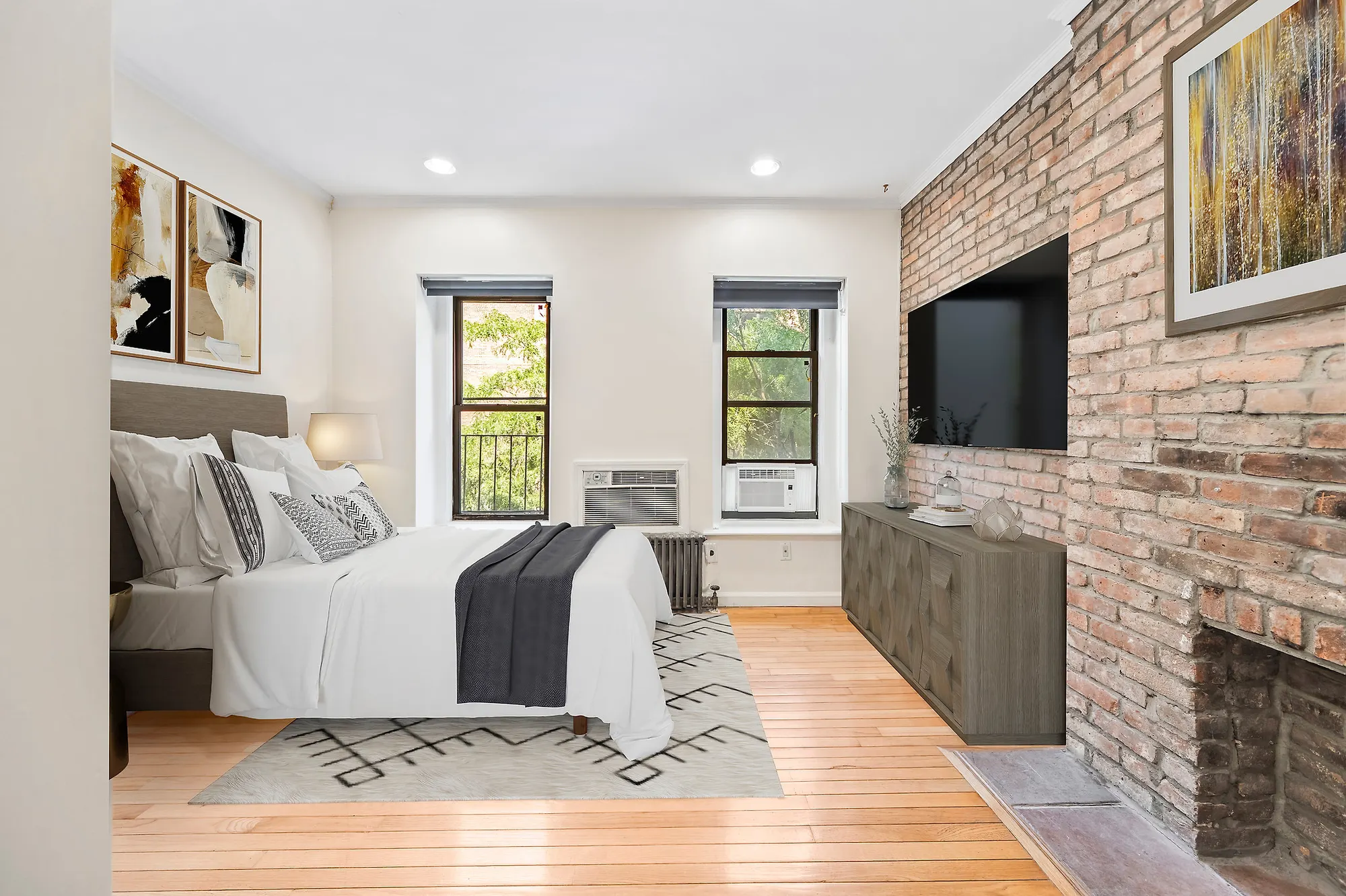 The same bedroom after virtual staging: a bed made up in white with a charcoal throw and patterned cushions against a gray upholstered headboard, a cream rug with a black geometric pattern, a wide dresser under a wall-mounted television on the brick, and framed abstract prints on the left wall. The windows, radiator and air conditioners are unchanged.