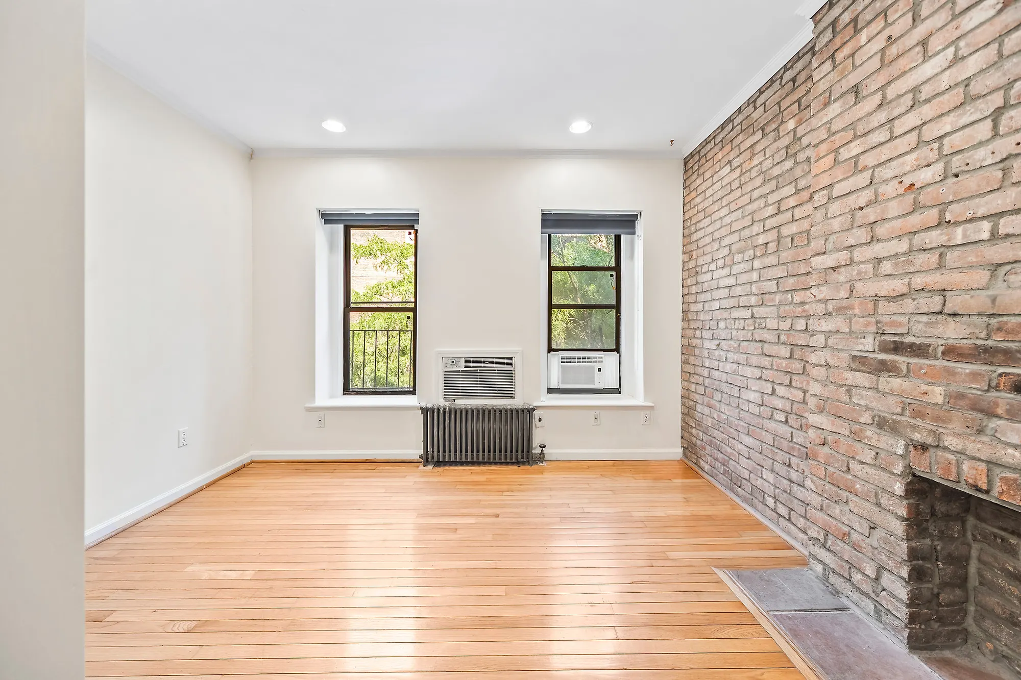 An empty bedroom in the same Upper East Side apartment: two windows, the left one with black security bars, a through-wall air conditioner between them above a cast-iron radiator and a second unit in the right window, an exposed brick wall on the right with a bricked fireplace opening, bare oak floors and white walls.