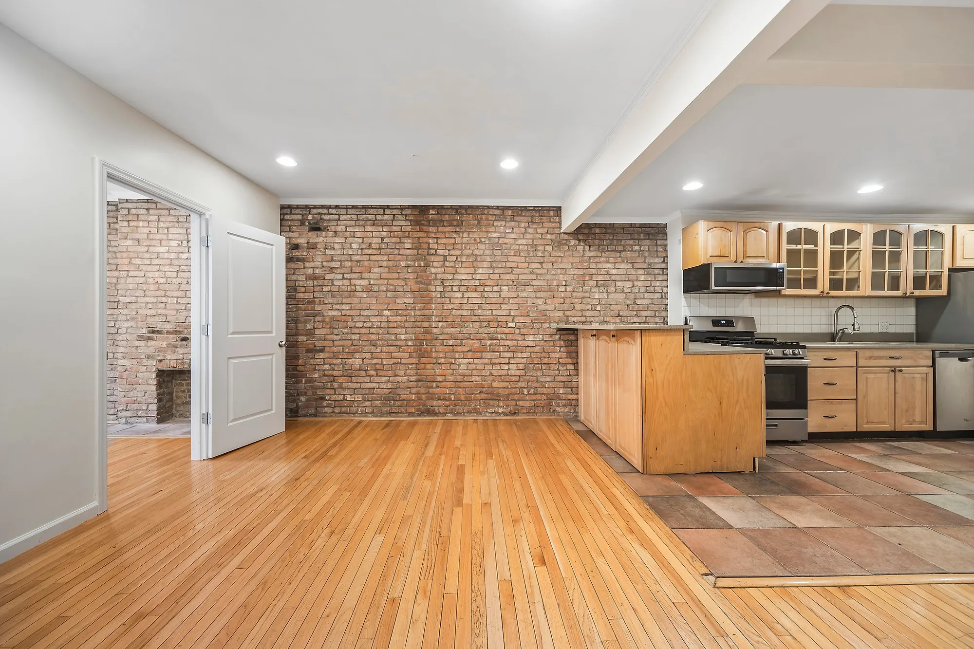 OpenDoors360 photo of the same Upper East Side apartment shot corner-on, an exposed brick wall meeting bare oak floors with a wood kitchen and a bricked fireplace opening beyond, the two walls and the floor line held in one frame with no furniture in it