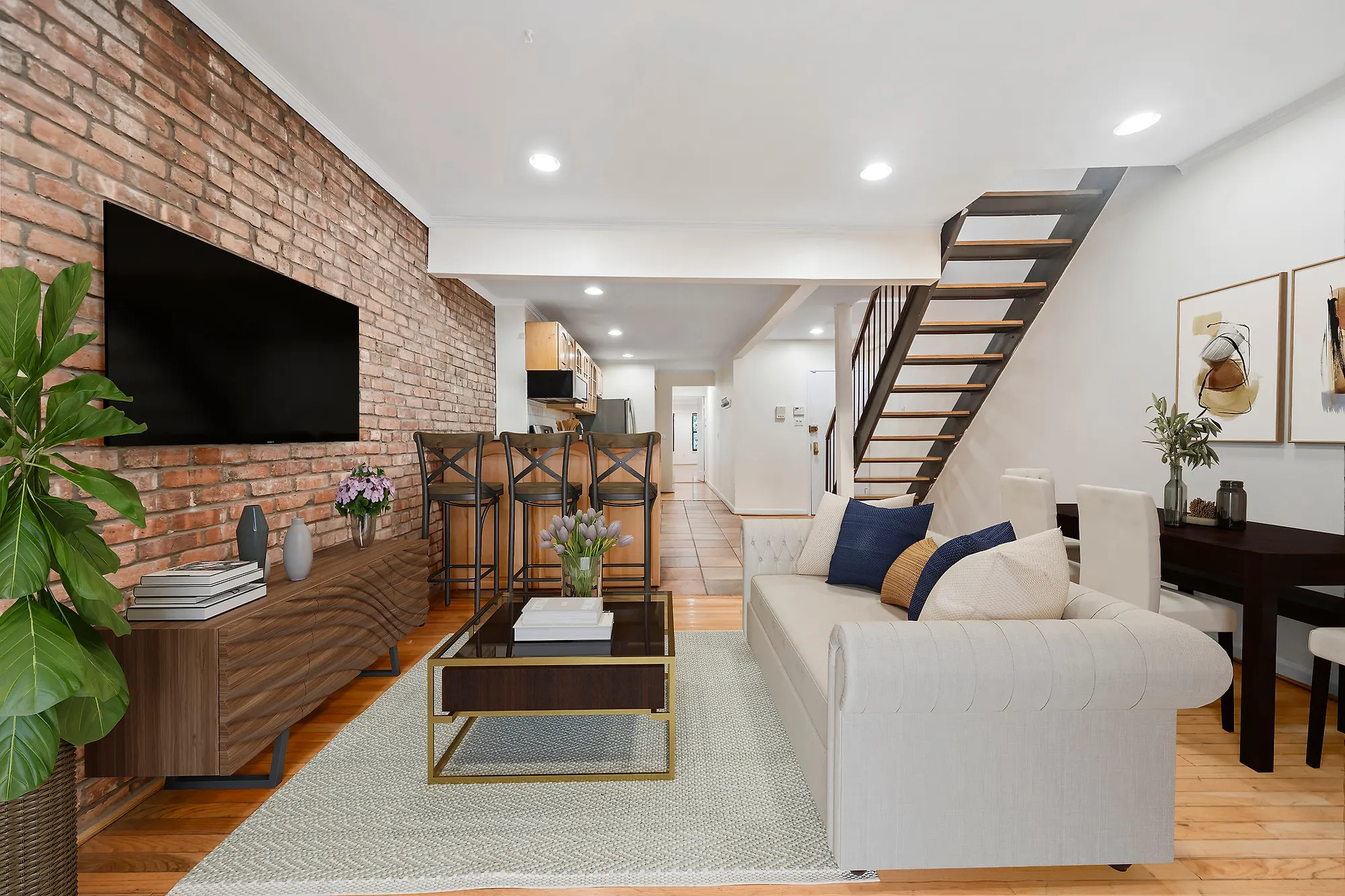 The same Upper East Side living room after virtual staging: a cream sofa with navy and tan pillows on a woven rug, a low walnut console under a wall-mounted television on the brick, a glass and brass coffee table, three counter stools at the kitchen bar, a fiddle-leaf plant in the foreground and framed prints on the right-hand wall.