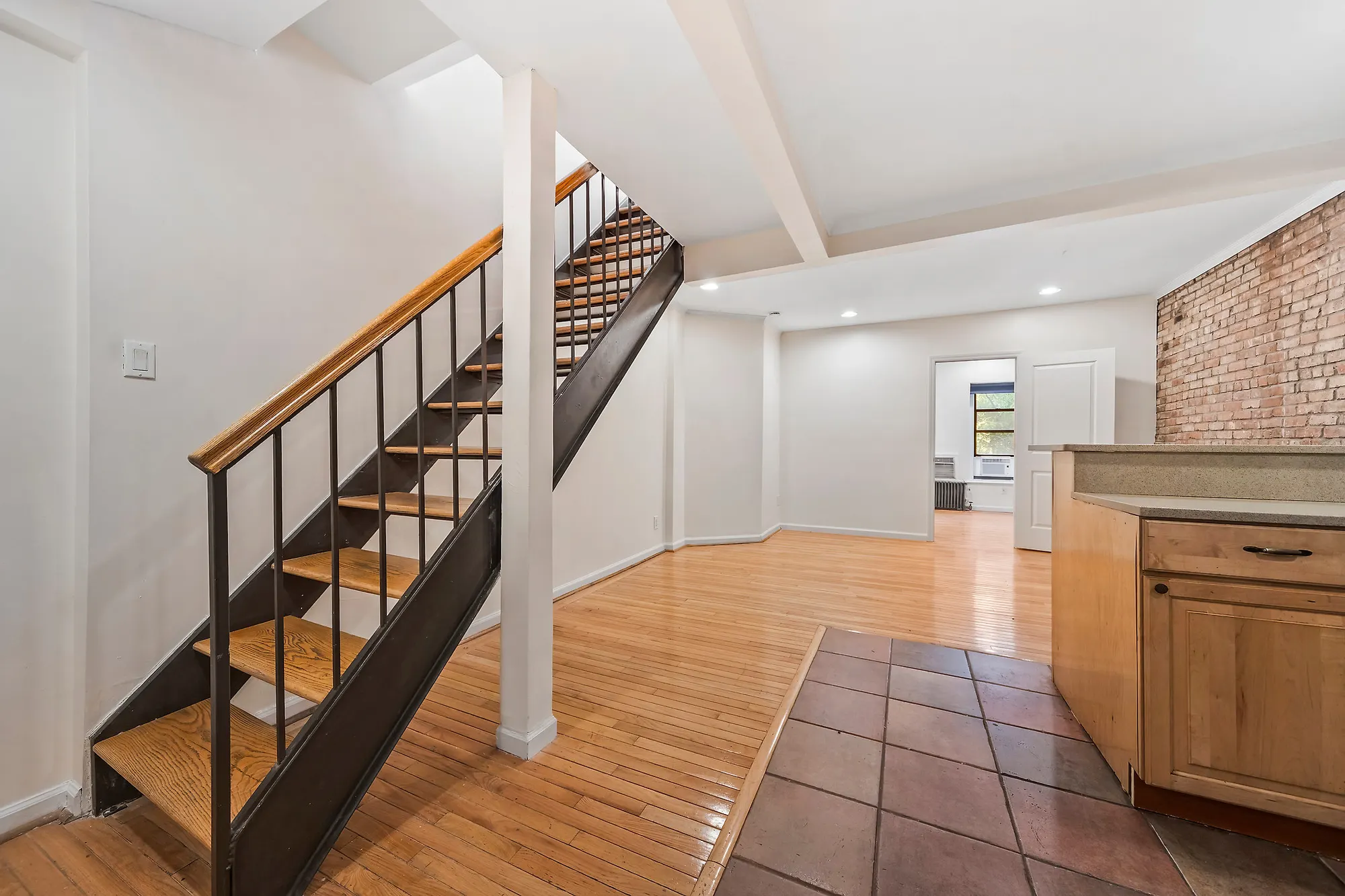 Entry of an Upper East Side duplex apartment with an open wood and black metal staircase, oak floors meeting a terracotta tile kitchen floor, a wood breakfast bar and exposed brick at the far wall
