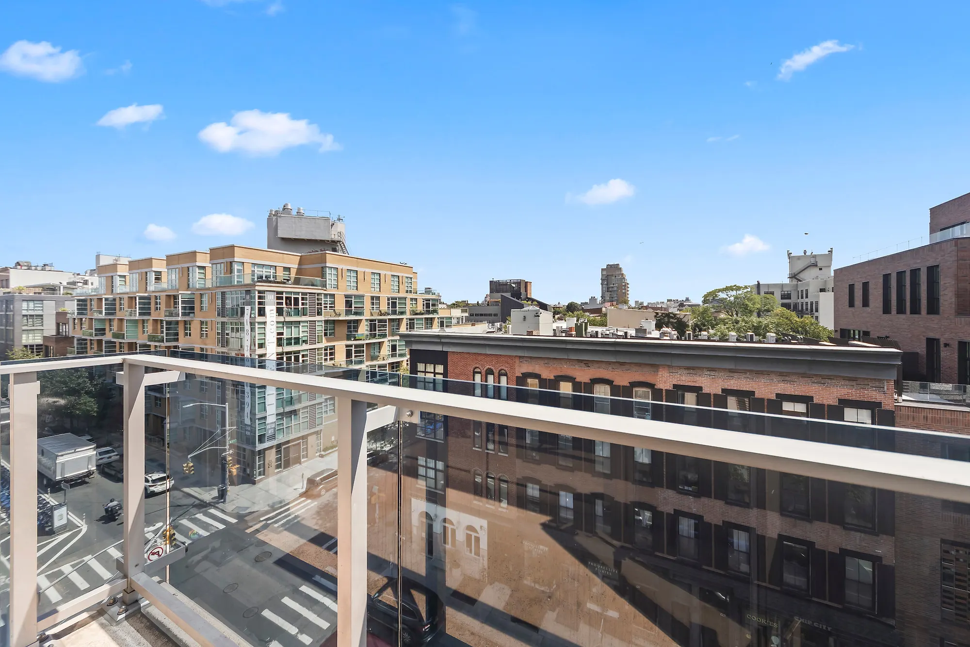 View from a Williamsburg balcony over North 7th Street, a low brick warehouse building opposite, a terraced apartment block beyond it and a clear blue sky