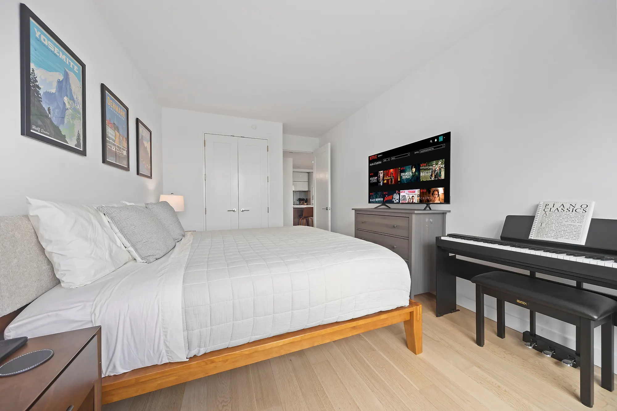 Bedroom in a Williamsburg condominium with a white bed on a wood frame, framed national park posters above it, a dresser under a wall-mounted television and an upright digital piano with sheet music open