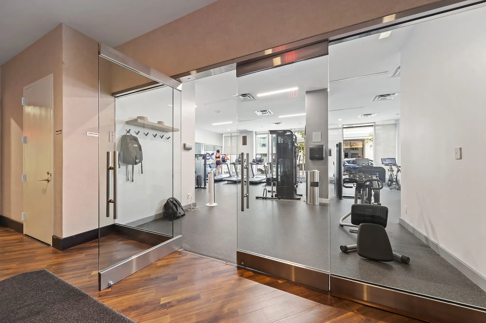 Glass-walled residents gym in a Williamsburg building, weight machines and cardio equipment behind mirrored walls, seen from the carpeted corridor outside