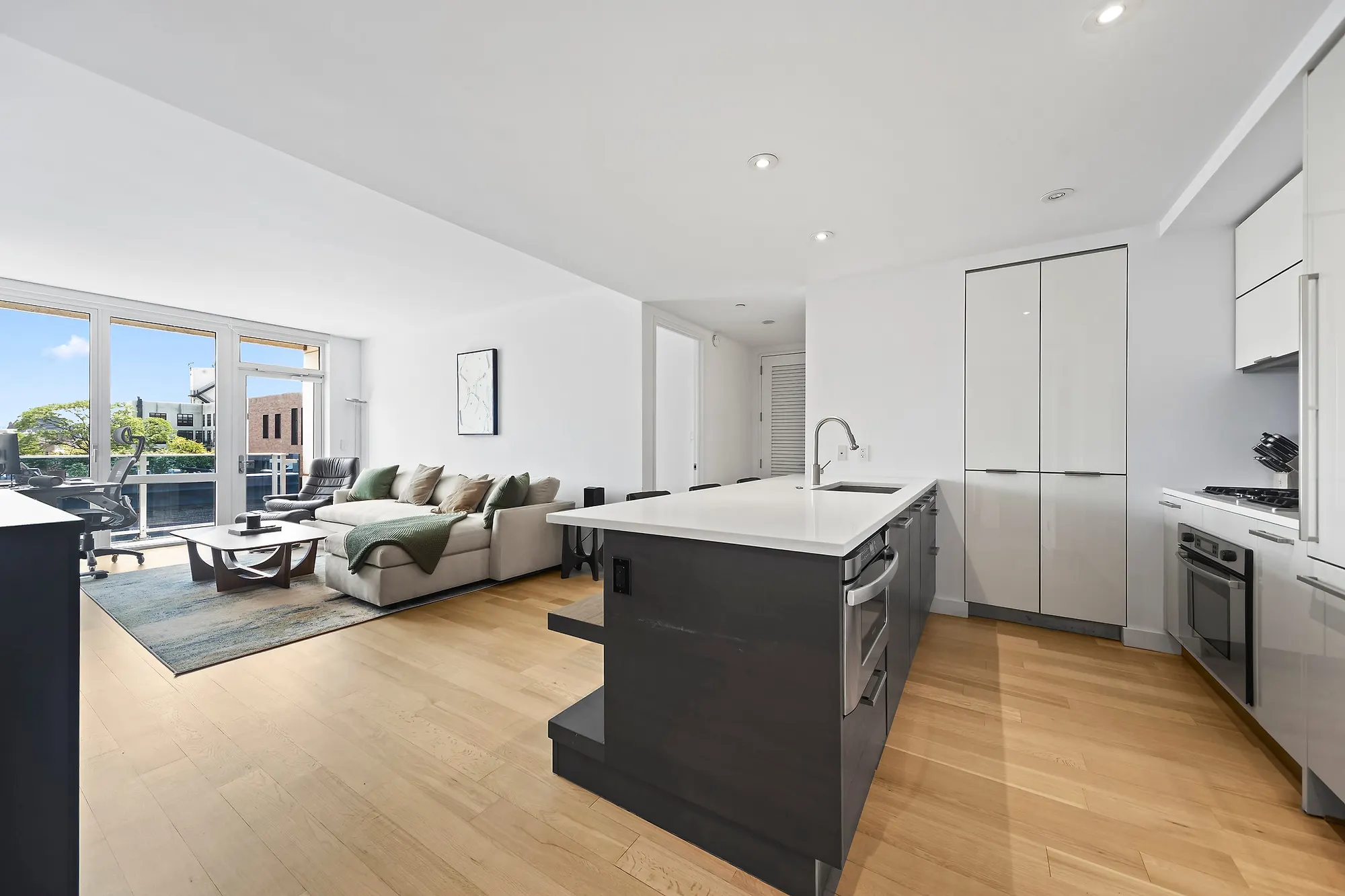 Open living and kitchen in a Williamsburg condominium with a white quartz island over a dark wood base, integrated panel appliances, pale oak floors and a sliding glass door to a balcony over the street
