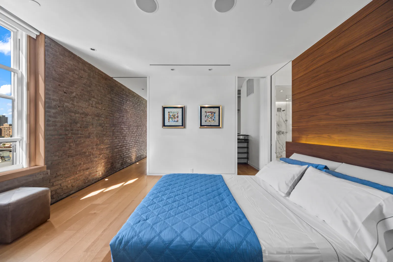 Bed with a blue quilt facing an exposed brick wall and a tall window in a SoHo loft bedroom, by OpenDoors360 in New York City