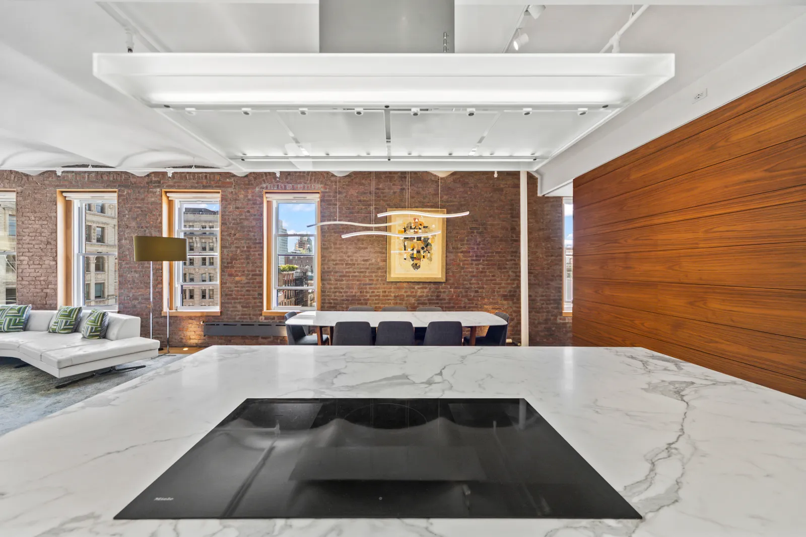 Black glass cooktop set into a marble island, looking across to the dining table and brick-framed windows of a Manhattan loft, by OpenDoors360
