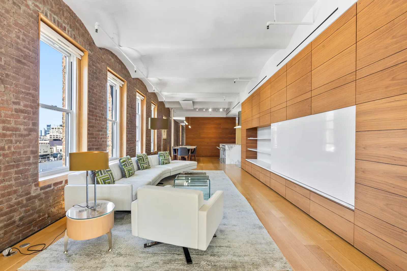 Long loft living room with a walnut-paneled wall, a white sofa and brick window bays on Broadway in SoHo, by OpenDoors360