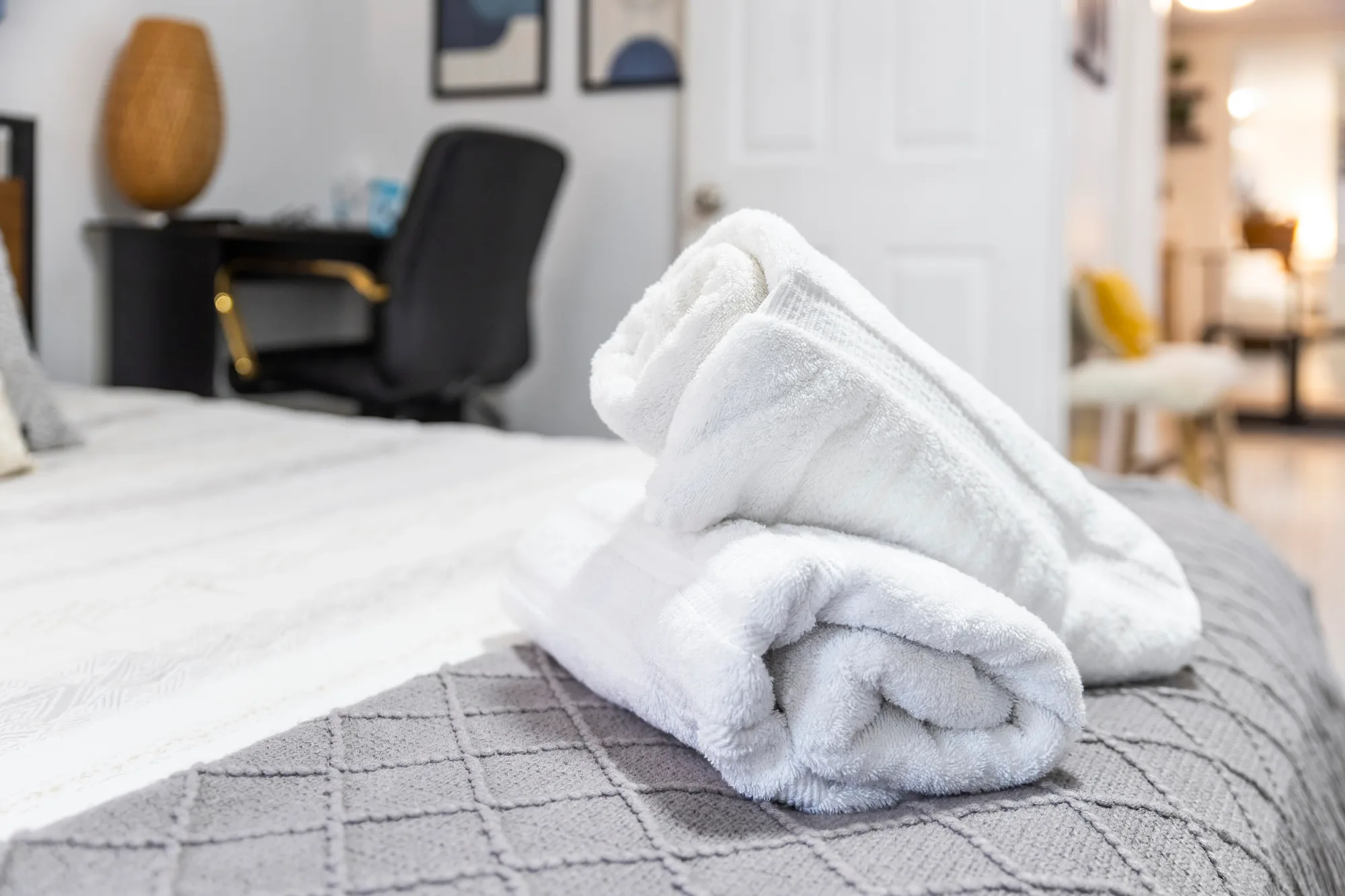 Close-up of rolled white spa towels staged on a bed with desk behind, by OpenDoors360
