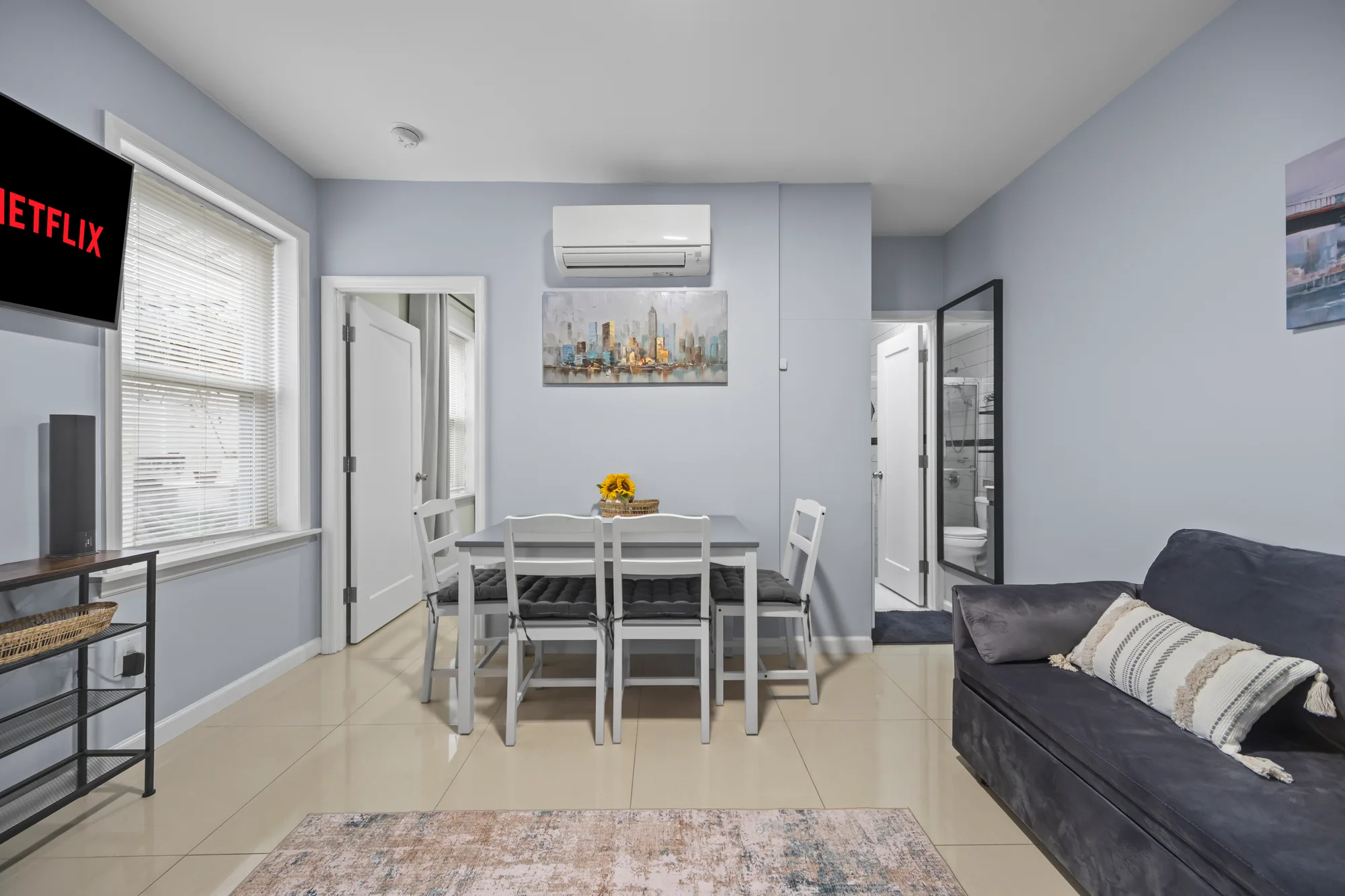 Gray-walled dining area with white table and chairs, wall-mounted TV, and city skyline art, by OpenDoors360