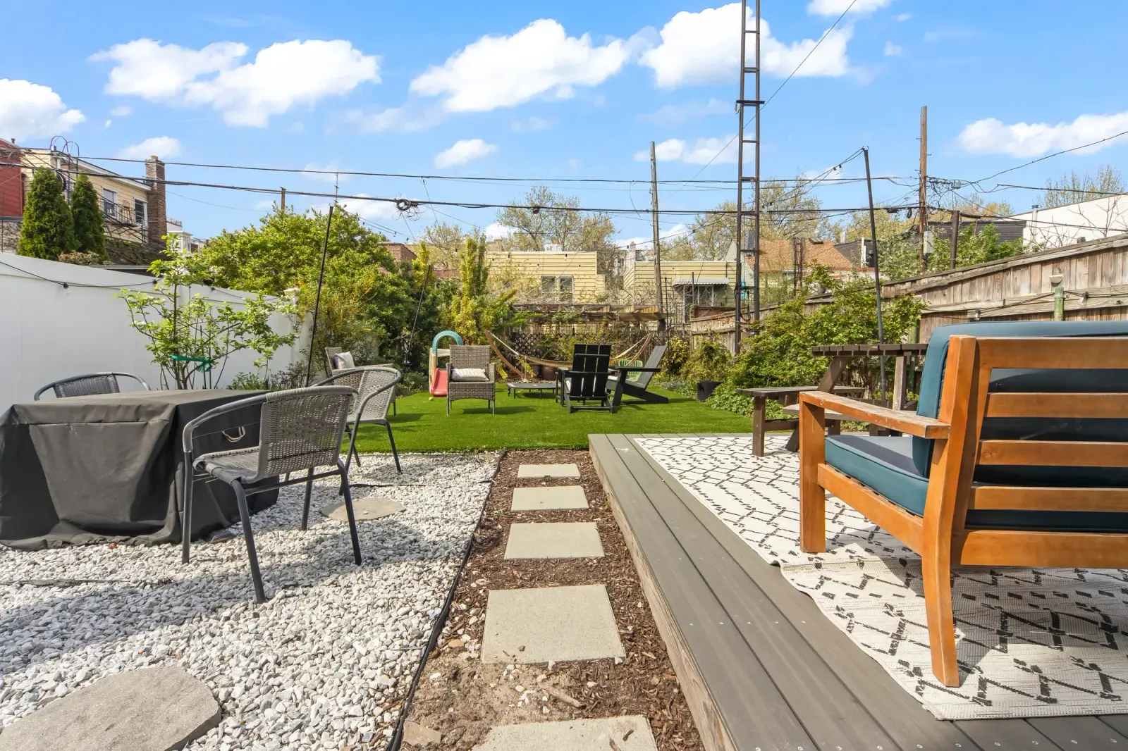 Sunny backyard with wood deck, patterned rug, gravel path, lawn, and patio seating, by OpenDoors360