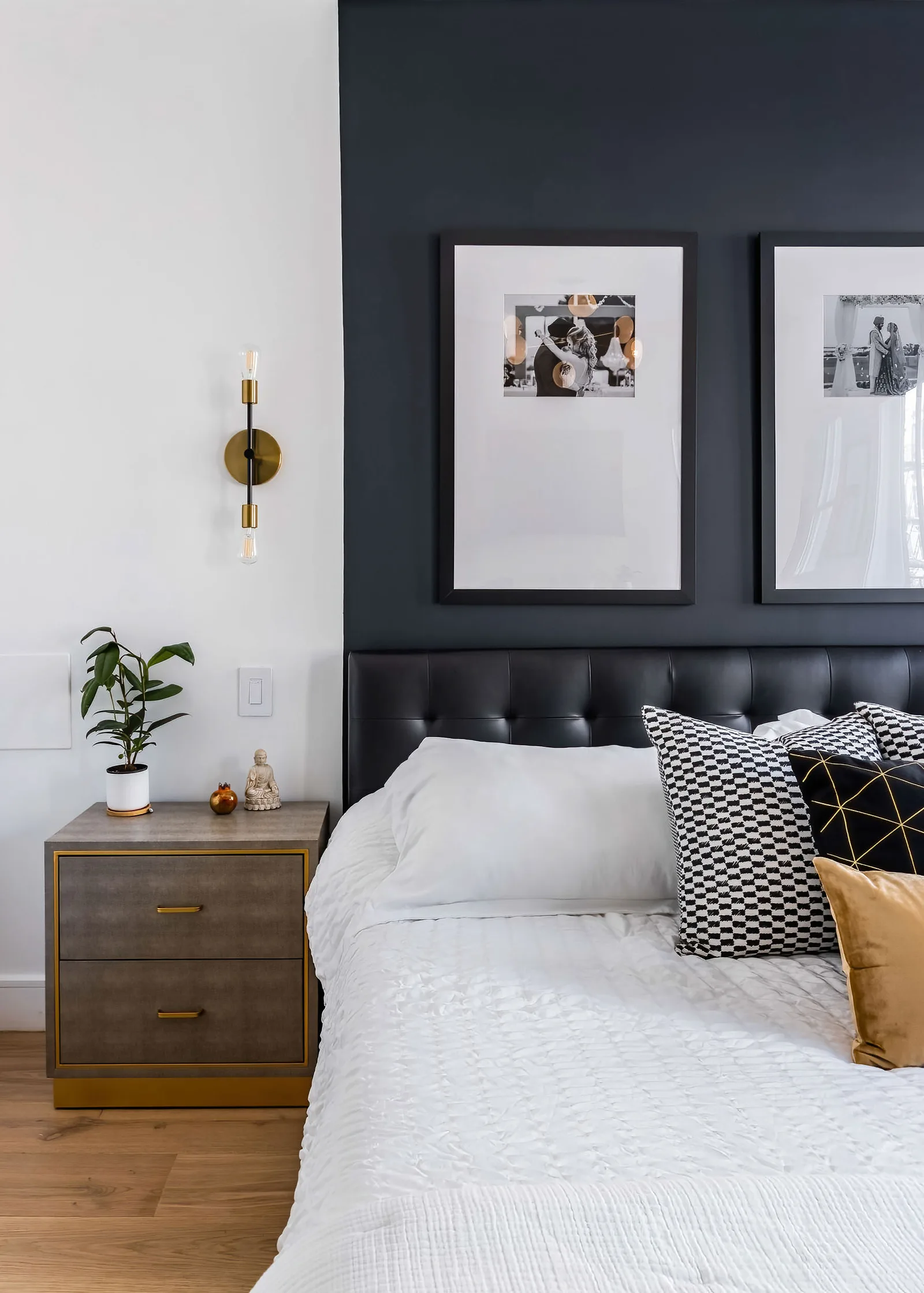 OpenDoors360 real estate photo of a bedroom with a black accent wall, two framed black and white wedding prints over a tufted black leather headboard, a brass sconce, a gray nightstand with a plant and a small Buddha figure, and a white bed with checkered and gold pillows, New York City