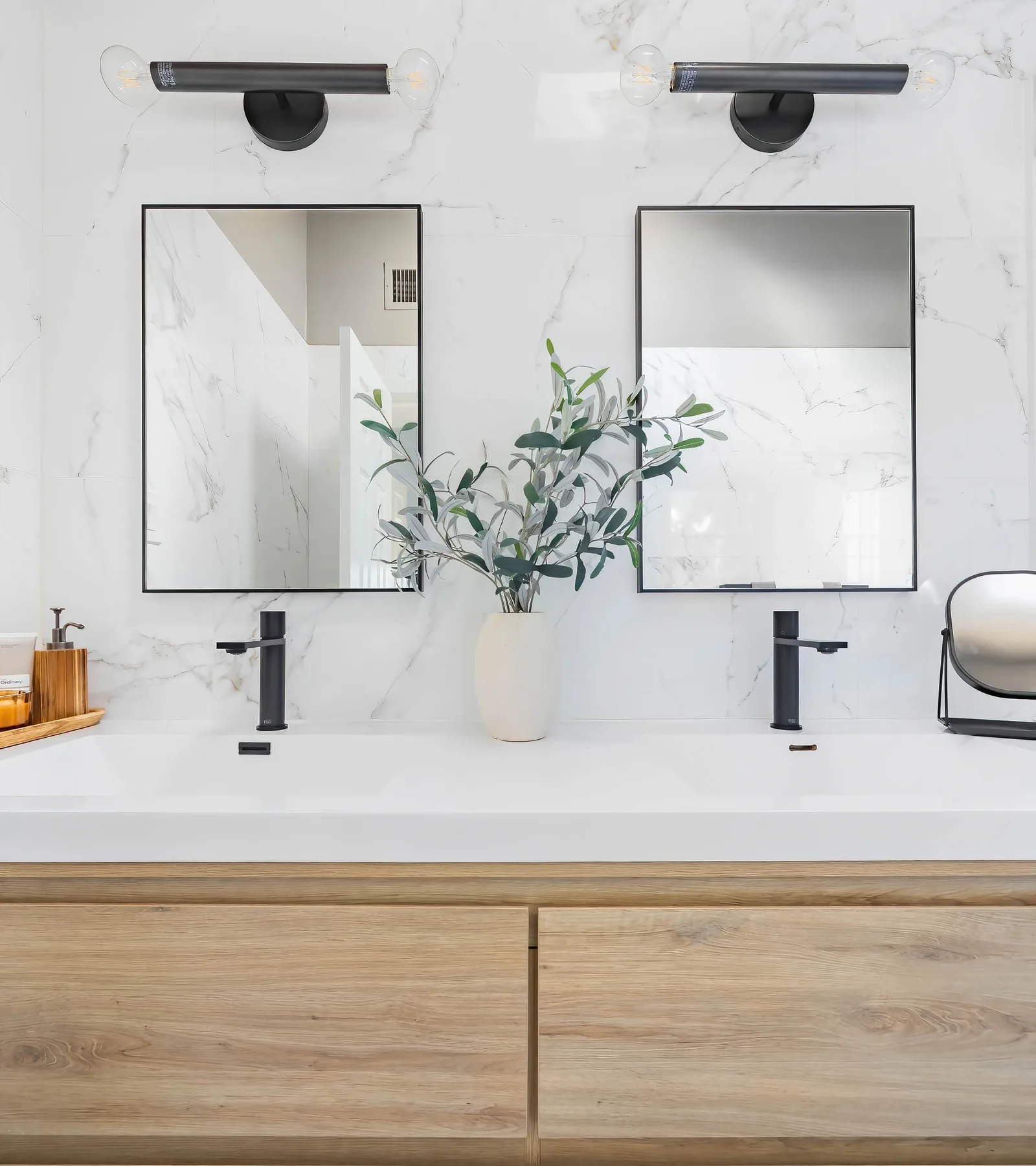 Double-sink bathroom vanity with marble walls, black framed mirrors, wood cabinetry, and olive plant, by OpenDoors360