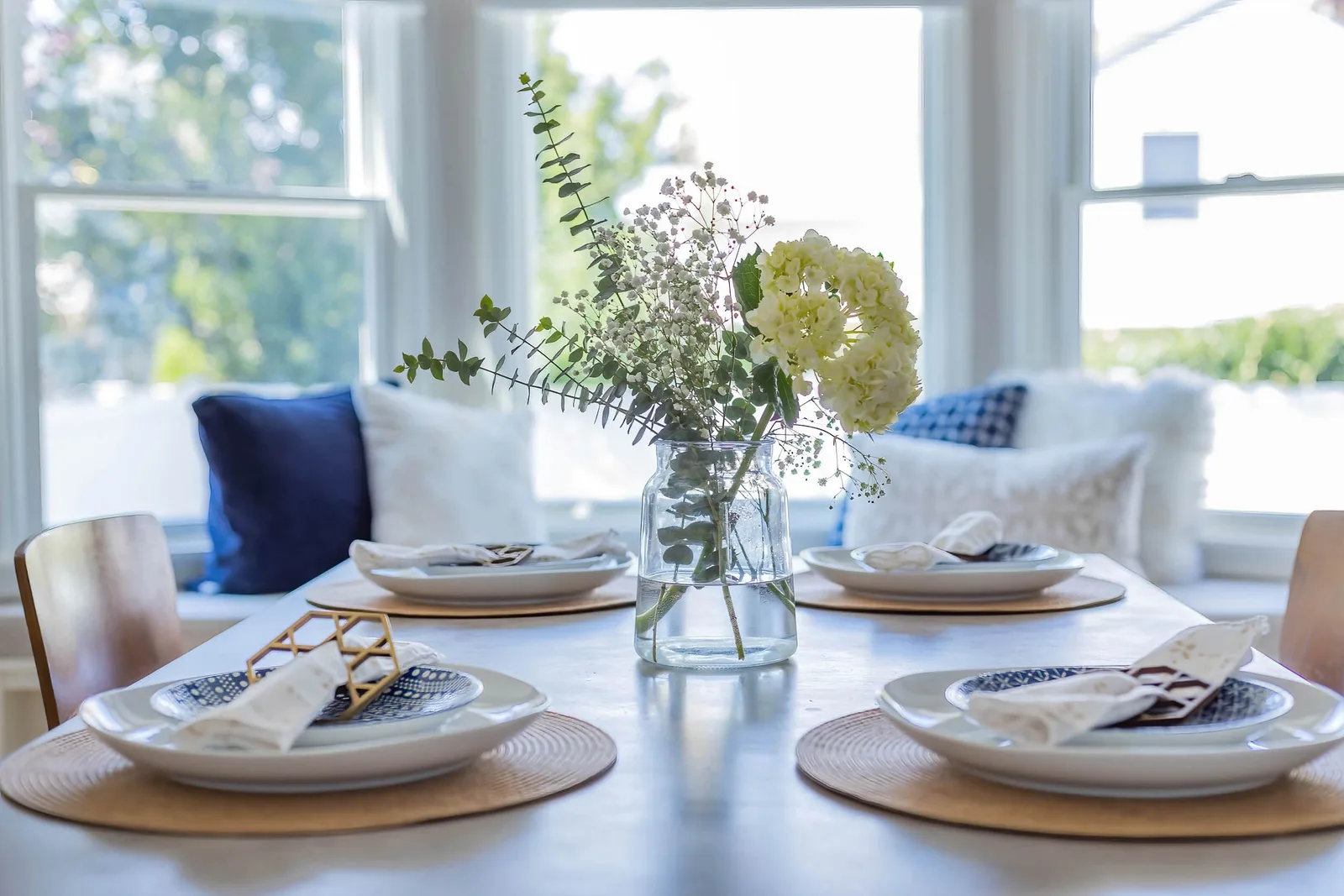 Dining table detail with white hydrangea bouquet, navy place settings, and woven placemats by bright windows, by OpenDoors360