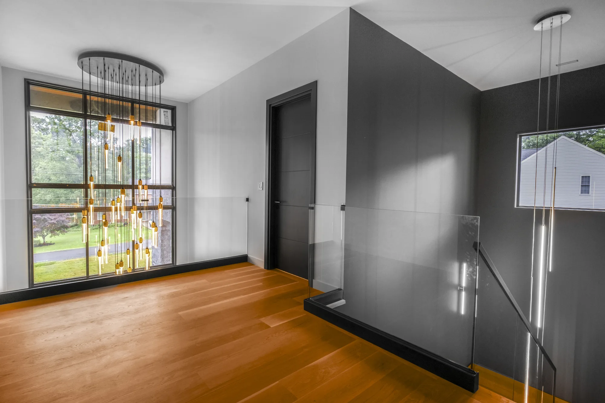Landing with cascading bead chandelier, dark accent wall, large window, and glass stair railing, by OpenDoors360