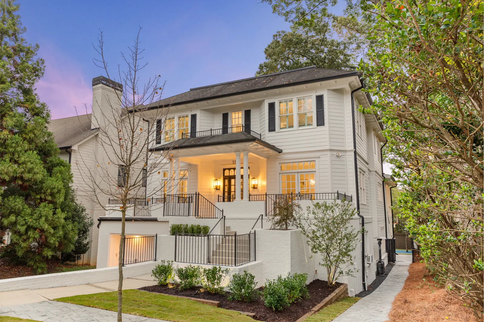 White colonial house at dusk from the front corner, porch lights on and landscaped beds along the walk, twilight real estate photography by OpenDoors360