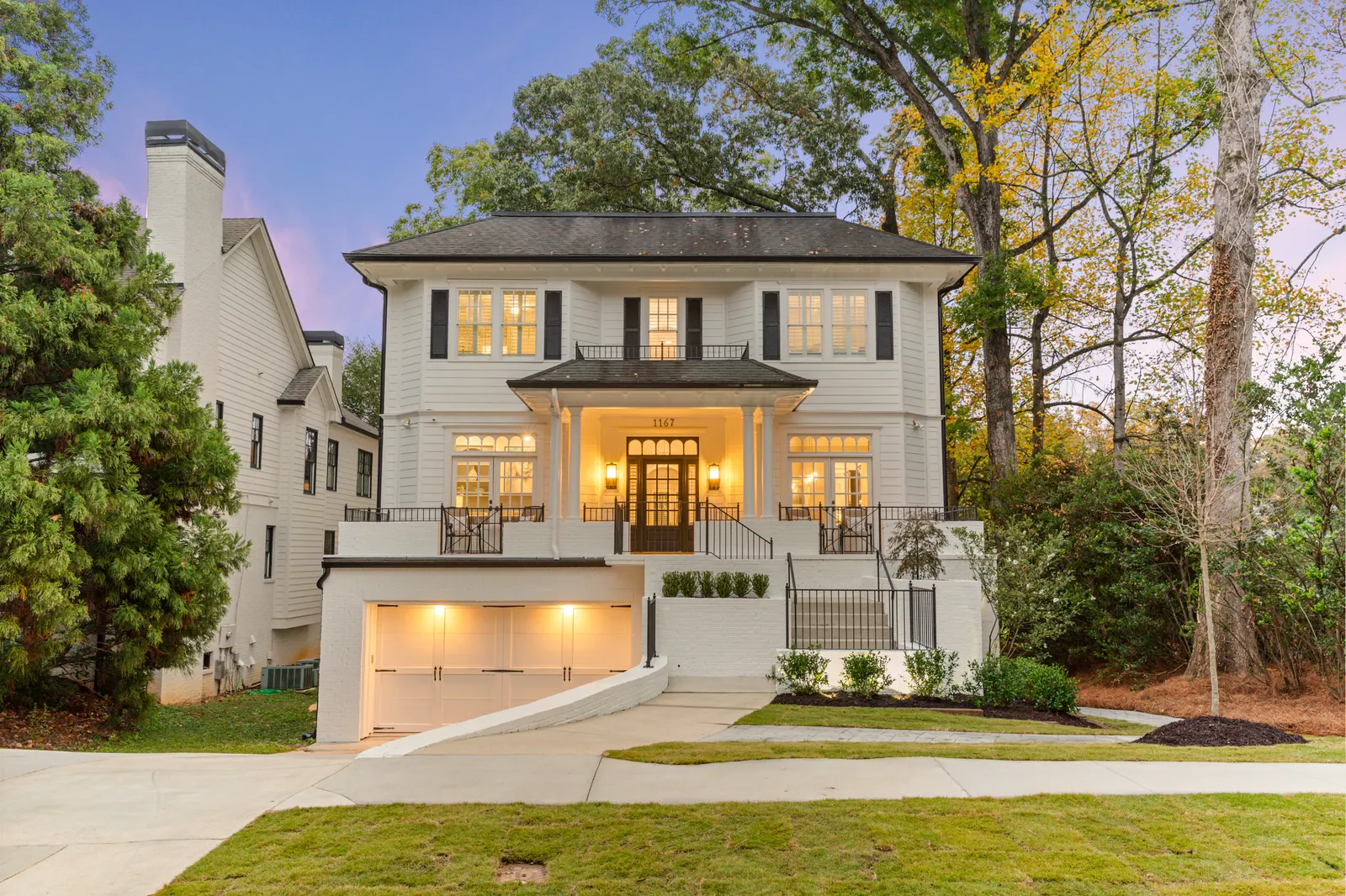 White colonial house photographed at dusk with every window lit, a covered front porch and a two-car garage under a violet sky, twilight real estate photography by OpenDoors360