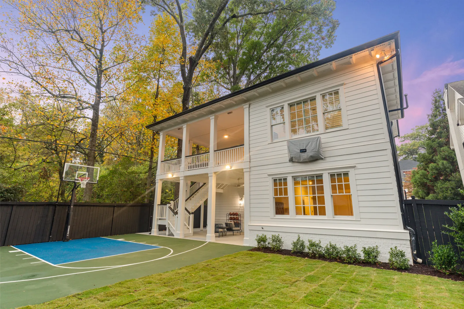 Rear of a white colonial house at dusk with a lit two-level porch and a backyard sport court, twilight real estate photography by OpenDoors360