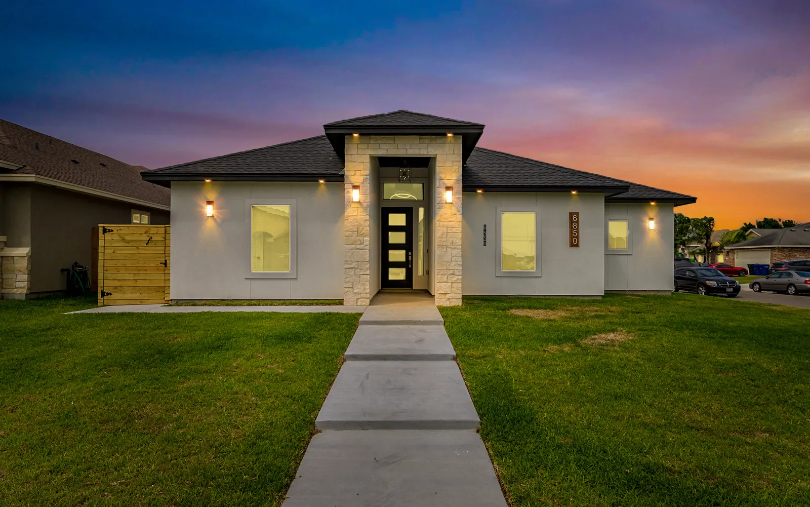 Single-story modern house at dusk with a stone-clad entry, lit wall sconces and a concrete walk across the lawn, twilight real estate photography by OpenDoors360