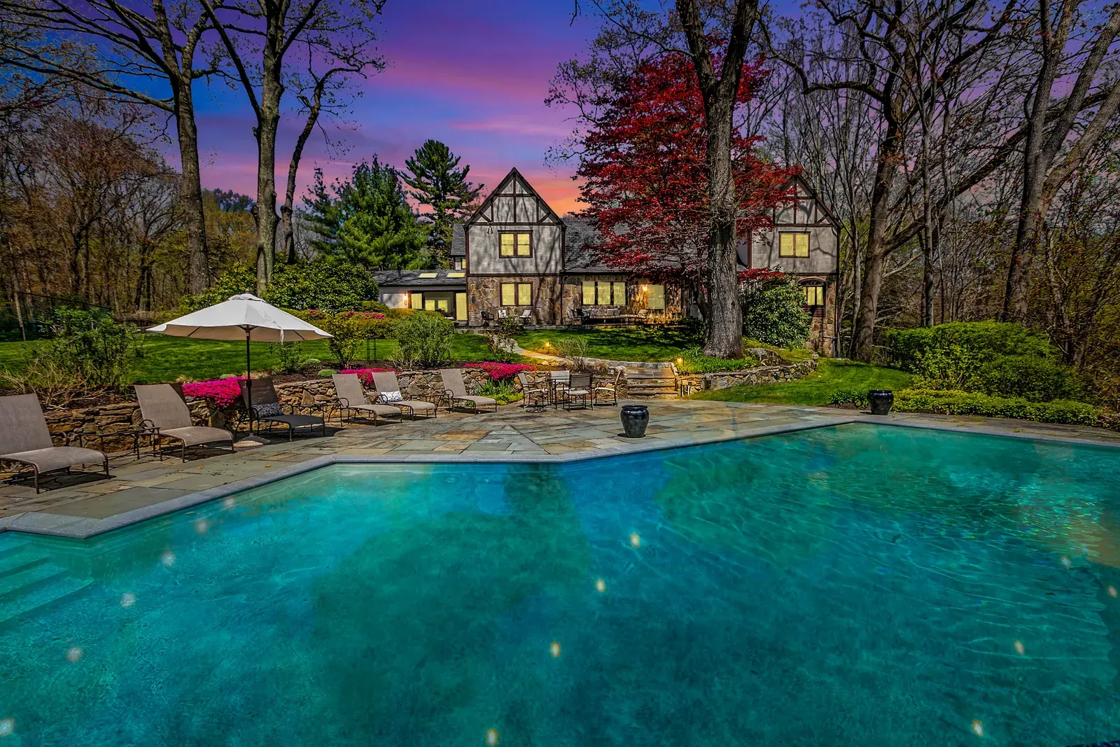 Tudor house at dusk above a lit swimming pool and stone patio with lounge chairs under a pink sky, twilight real estate photography by OpenDoors360