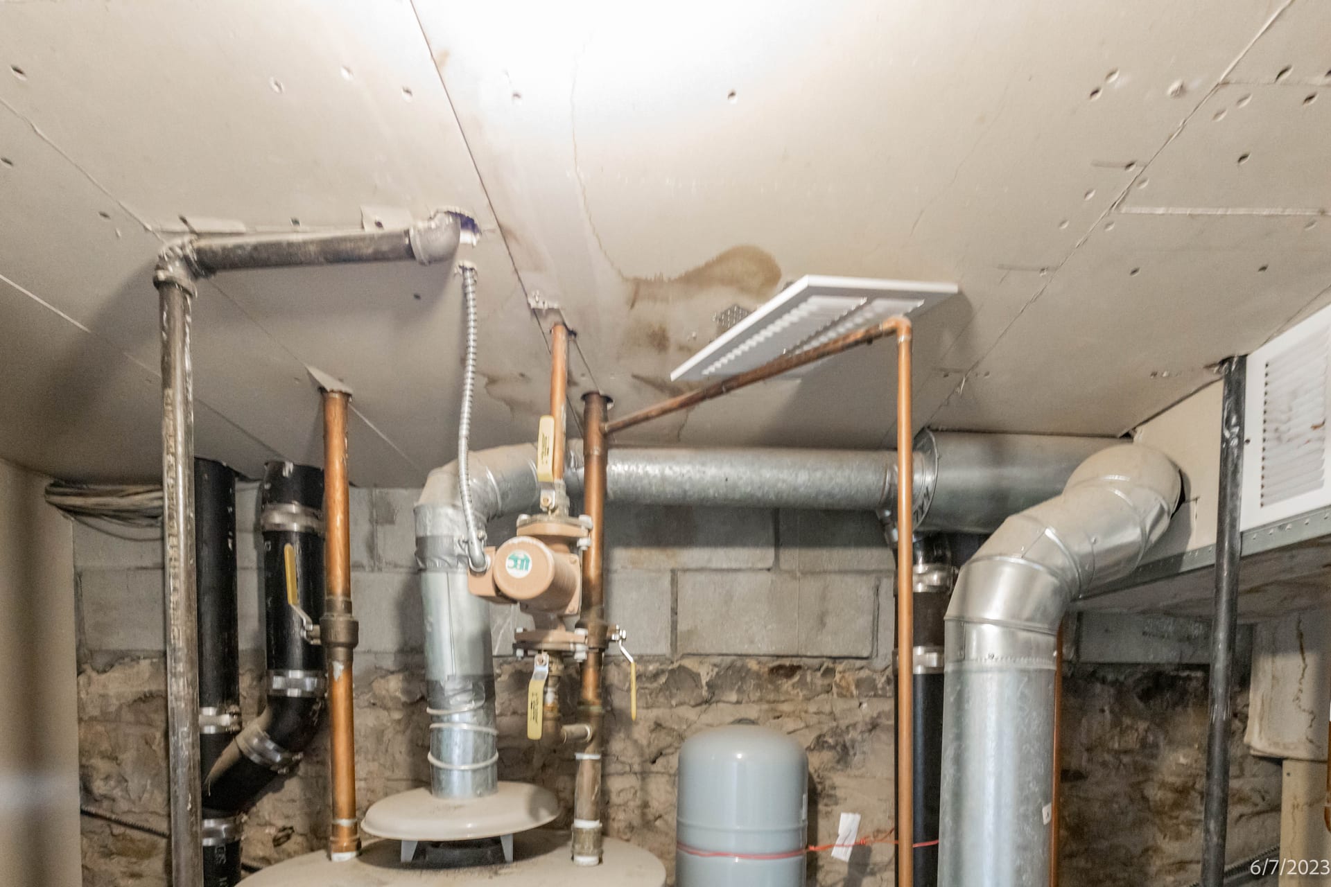 OpenDoors360 survey frame in a Brooklyn basement mechanical space, with cinder-block and rubble-stone walls, copper and black iron pipes, a brass backflow preventer, the top of a water heater, a gray expansion tank, galvanized ductwork, and a drywall ceiling carrying a brown water stain, a visible seam crack and a supply register